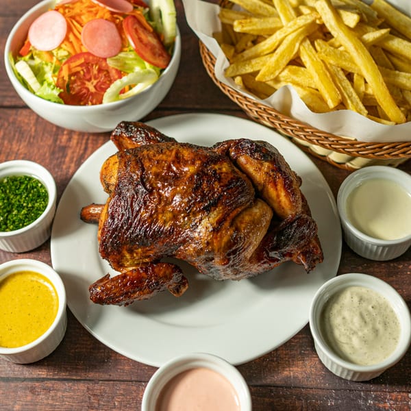 Whole roasted chicken on a white plate with fries, salad, and various dipping sauces.