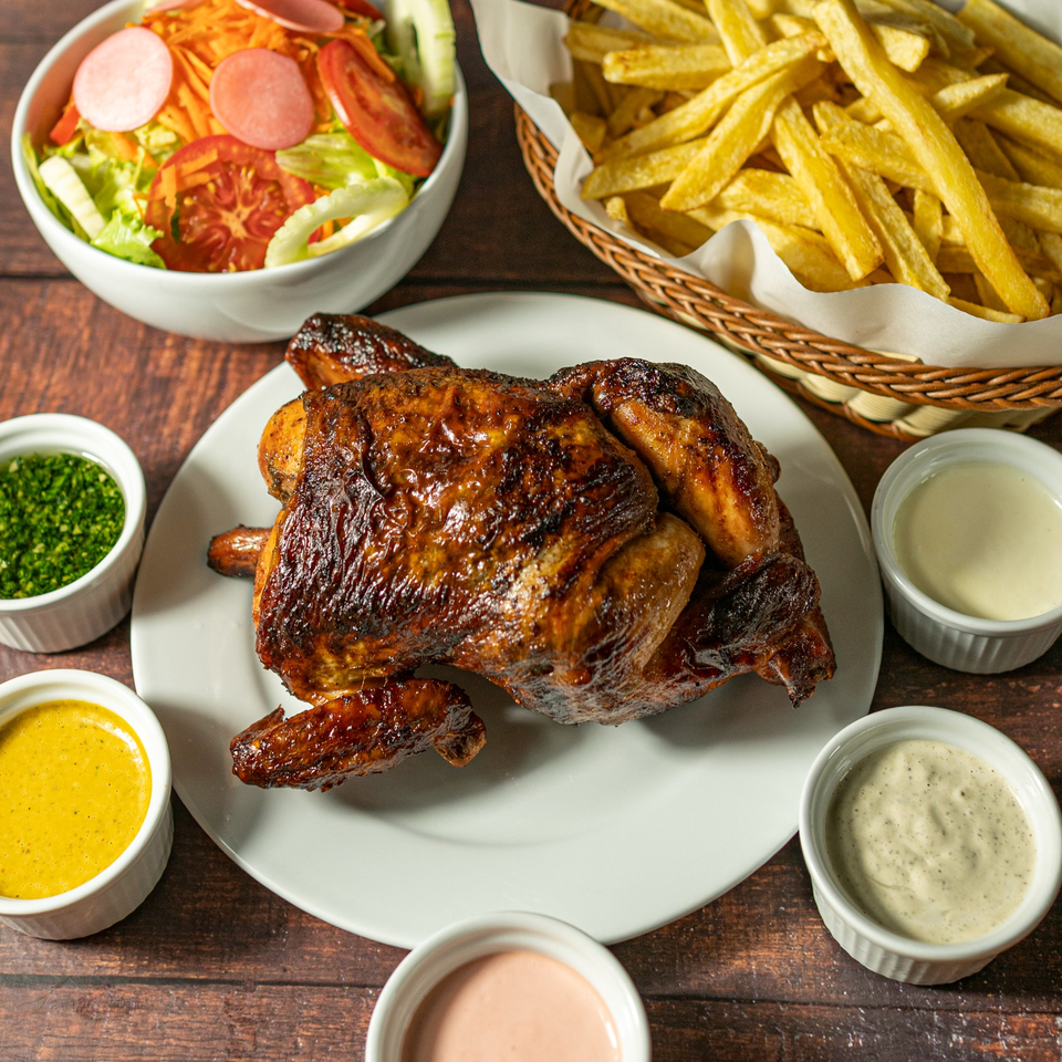 Whole roasted chicken on a white plate with fries, salad, and various dipping sauces.